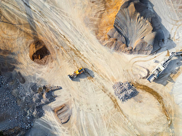 Aerial view of a gravel quarry with heavy machinery, large piles of aggregate, and dirt paths under bright sunlight, showing industrial activity.